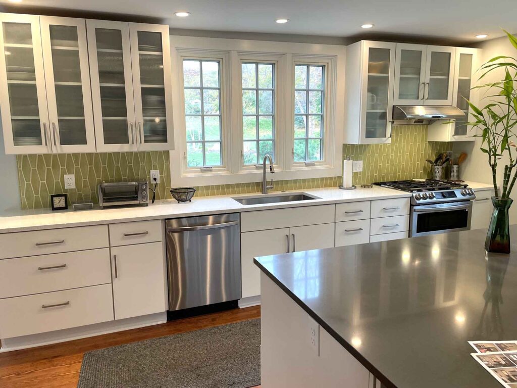 Kitchen renovation featuring white, glass-front upper cabinets, quartz countertops, stainless-steel appliances, green tile backsplash, and a wood floor.